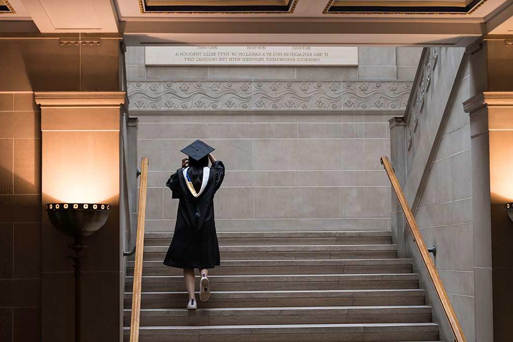 Student walks up stairs of Rush Rhees Library in cap and gown